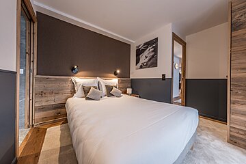 A cozy, modern bedroom with a white bed, rustic wooden headboard, dark accent walls, and illuminated wall sconces. Doors lead elsewhere.