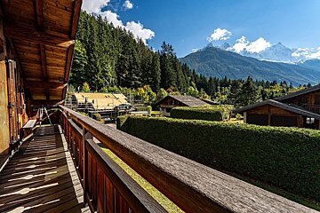 Scenic view from a wooden balcony: alpine chalets, lush forest, and majestic snow-capped mountains under a clear blue sky.