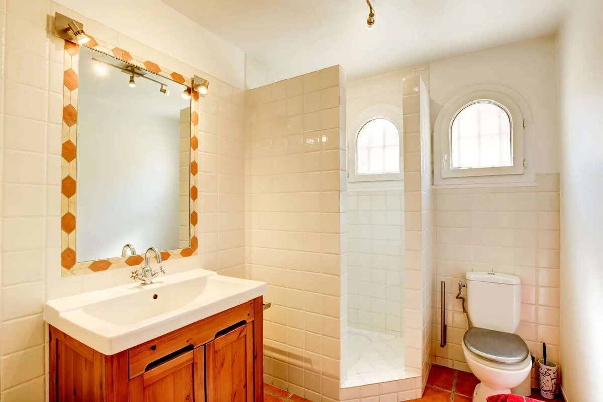 A bright bathroom with white tiled walls and terracotta floor, featuring a wooden vanity, mirror, walk-in shower with arched windows, and a toilet.