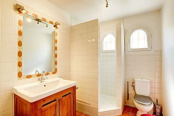 A bright bathroom with white tiled walls and terracotta floor, featuring a wooden vanity, mirror, walk-in shower with arched windows, and a toilet.