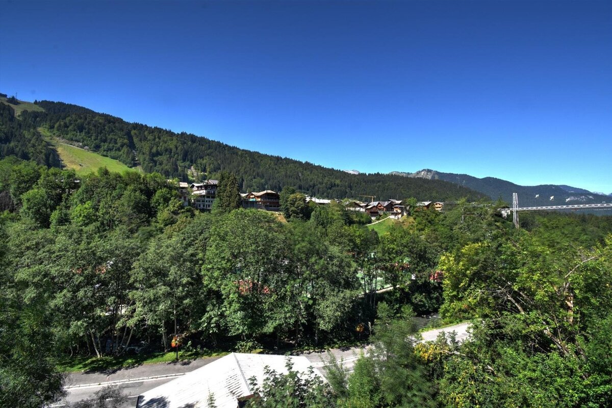 Scenic mountain landscape featuring lush green forests, alpine houses, a long suspension bridge, and a clear blue sky.