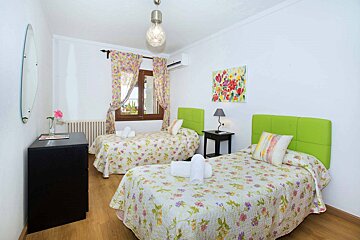A bright twin bedroom features white walls, lime green headboards, floral bedspreads and curtains, a wooden floor, and a colorful abstract painting.