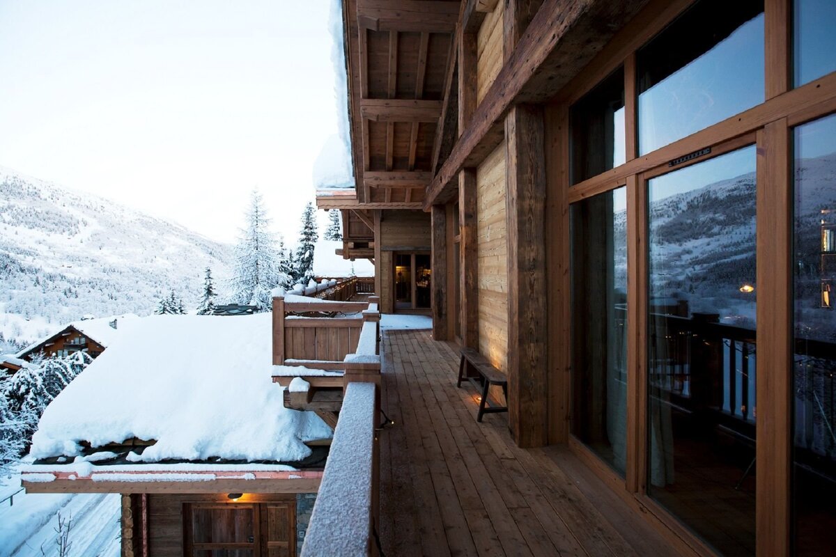 A rustic wooden chalet balcony covered in fresh snow, offering a scenic view of snow-capped mountains and a winter village.