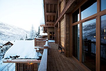 A rustic wooden chalet balcony covered in fresh snow, offering a scenic view of snow-capped mountains and a winter village.