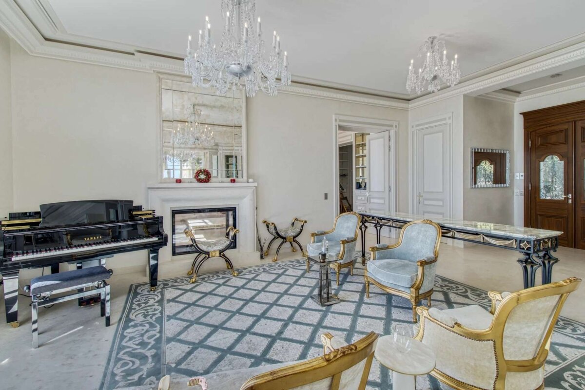 An opulent living room with a grand piano, fireplace, large mirror, crystal chandeliers, and classic ornate furniture on a patterned rug.