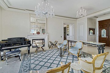 An opulent living room with a grand piano, fireplace, large mirror, crystal chandeliers, and classic ornate furniture on a patterned rug.