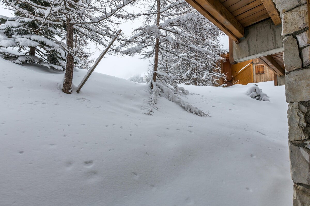 A winter scene with a deep snow-covered slope, frosted trees, and a wooden chalet partially visible on the right.