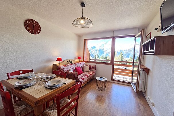 Cozy alpine apartment interior with a set dining table, red chairs, patterned sofa, and balcony views of snowy mountain peaks.