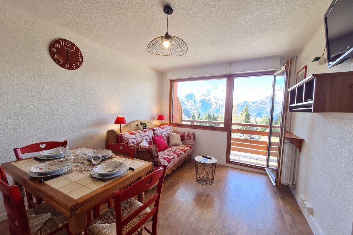 Cozy alpine apartment interior with a set dining table, red chairs, patterned sofa, and balcony views of snowy mountain peaks.