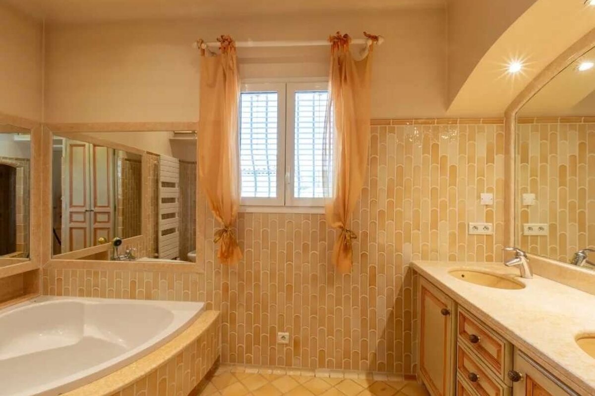 A warm-toned bathroom features a corner tub, double vanity, mirror, and window with orange curtains. Beige/yellow tiles cover walls and floor.