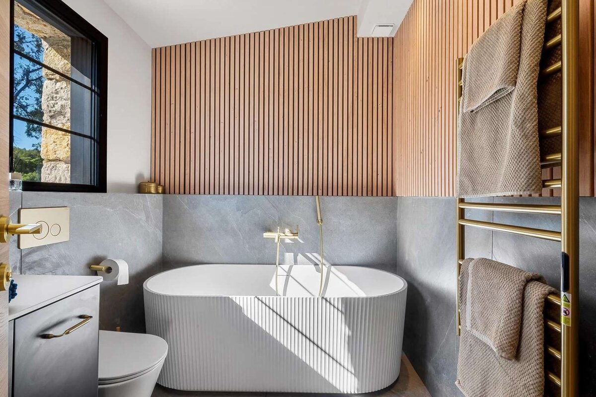 Elegant modern bathroom featuring a white fluted bathtub, light wood slat wall, grey stone tiles, and stylish gold fixtures, bathed in sunlight.