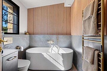 Elegant modern bathroom featuring a white fluted bathtub, light wood slat wall, grey stone tiles, and stylish gold fixtures, bathed in sunlight.