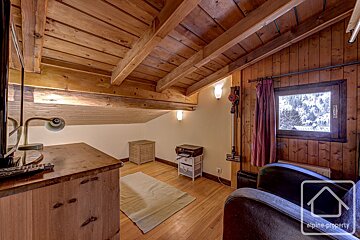 A cozy, wood-paneled attic room with exposed beams, a window overlooking a snowy landscape, and rustic furniture, including a desk and armchair.