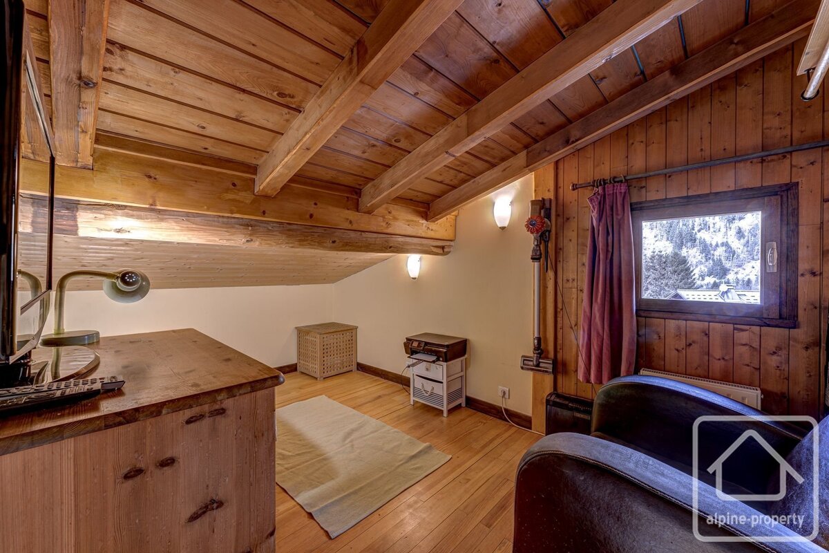 Cozy attic room with exposed wooden ceiling beams, wood paneled walls, a small desk, armchair, and a window overlooking snowy trees.