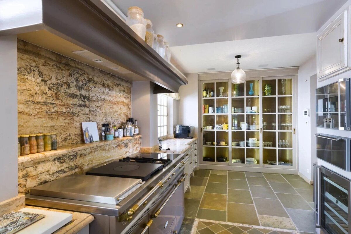 A spacious, elegant kitchen featuring a large professional stove, stone-tiled floor, and a tall glass-front display cabinet.