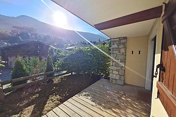 A sunny wooden patio with a stone pillar, overlooking a mountain village nestled in a valley with bright sun and green trees.
