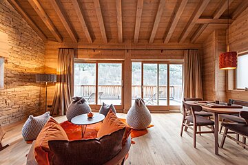A cozy, rustic chalet interior with exposed wooden beams, stone wall accents, beanbag seating, and large windows overlooking a scenic view.