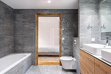 Modern gray tiled bathroom with a white bathtub, toilet, double sink vanity, and an open doorway to a bedroom.