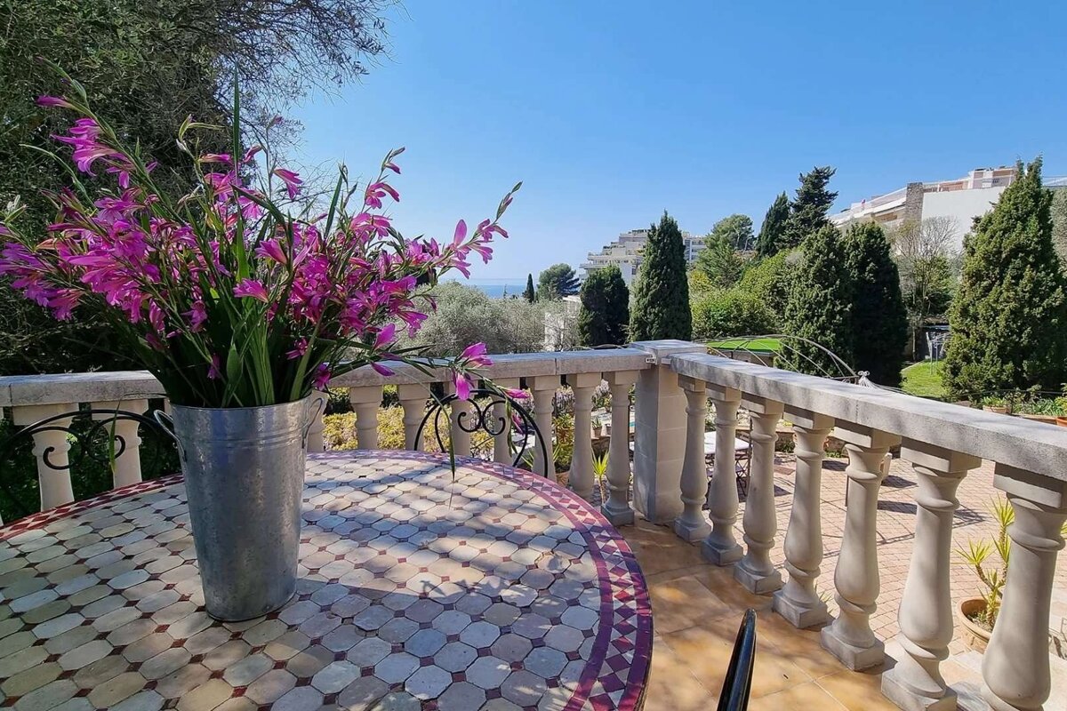 Sunny outdoor balcony with vibrant purple flowers on a mosaic table. Features a scenic view of green trees, distant buildings, and a clear blue sky.