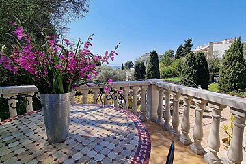 A vibrant bouquet of pink flowers sits on a tiled table on a sunny balcony, overlooking a lush landscape, buildings, and the distant blue sea under a clear sky.