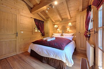 A cozy, rustic chalet bedroom with light wooden walls, exposed beams on a slanted ceiling, a bed with white sheets and red bedding, and red curtains.
