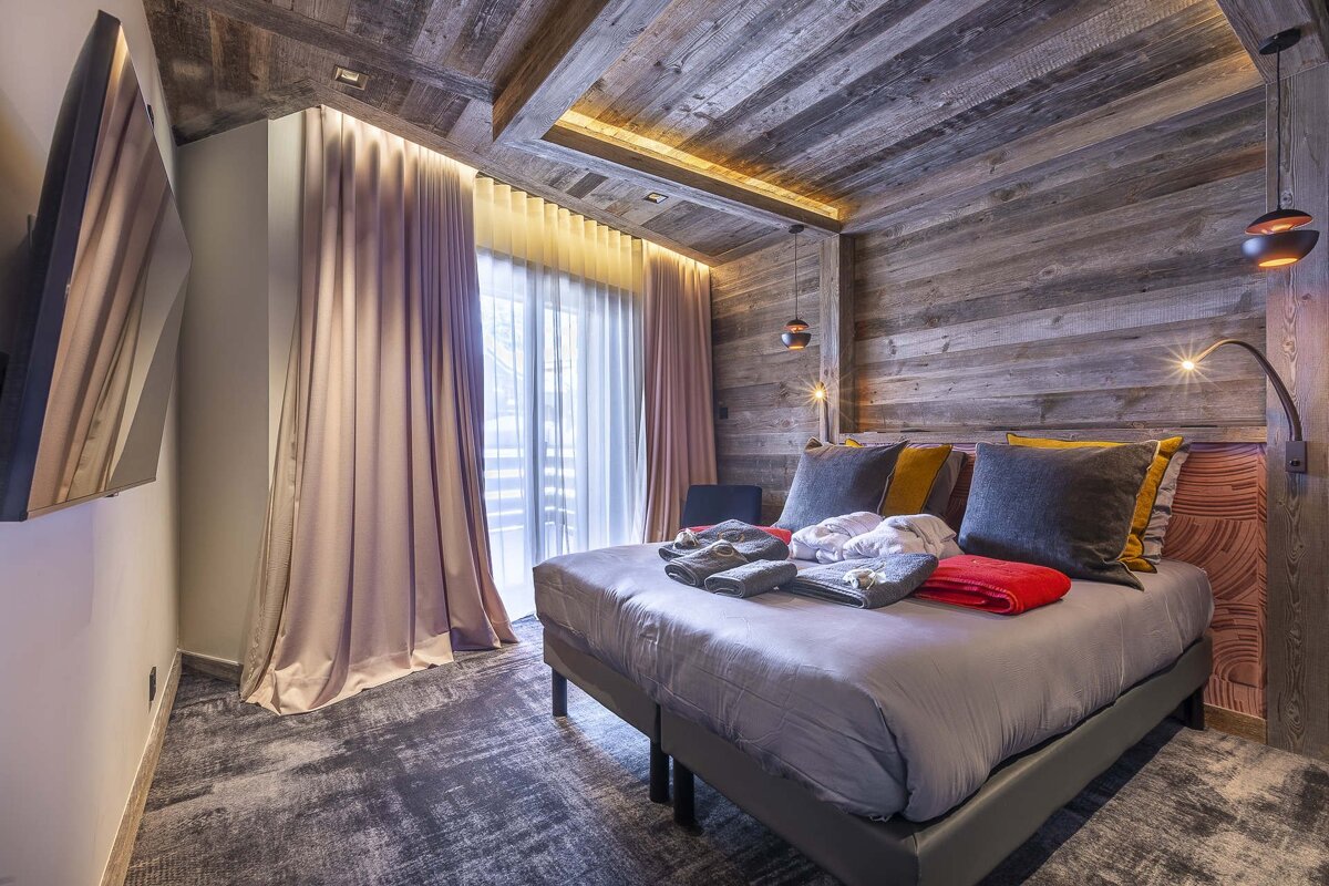 A cozy, modern bedroom with wood paneled walls/ceiling, a large bed with colorful pillows, a balcony, and contemporary lighting.