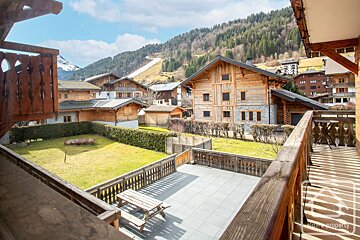 View from a balcony of an alpine village with wooden chalets, a green garden, and a distant ski slope on a forested mountain under a blue sky.