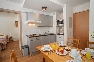 A compact apartment interior with a modern gray kitchenette, a dining table set for breakfast, and a partial view of an adjacent bedroom.