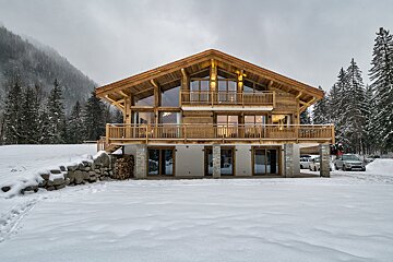 A grand wooden chalet stands in a snowy mountain landscape, surrounded by pine trees under a cloudy winter sky. The ground is covered in white snow.