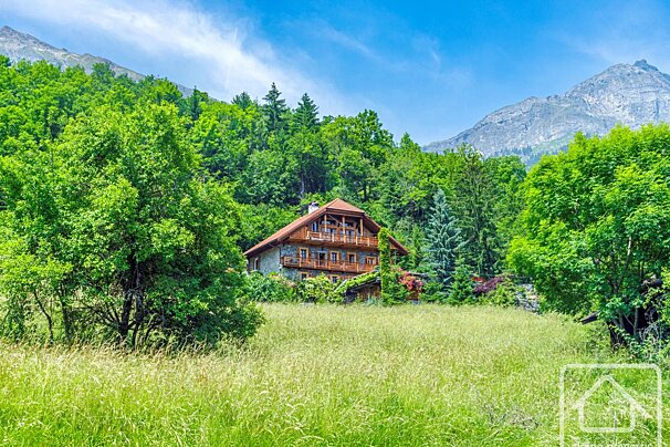 A rustic wooden chalet nestled in a lush green mountain forest with a grassy meadow in the foreground under a blue sky.
