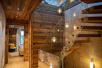 A luxurious wooden chalet interior featuring a modern floating staircase with glass railings and hanging globe lights, leading to an open doorway.