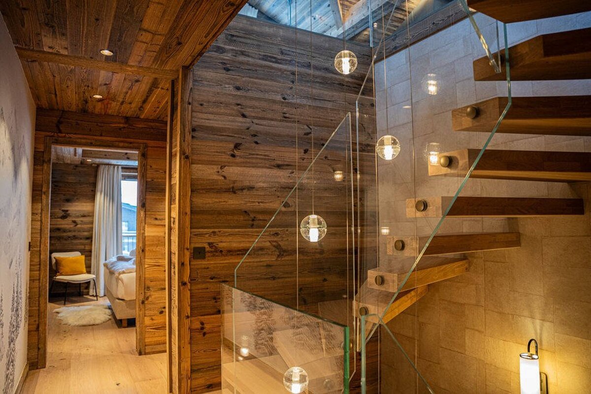 Modern chalet interior featuring rustic wood walls, a sleek glass staircase with floating lights, and a peek into a cozy bedroom.
