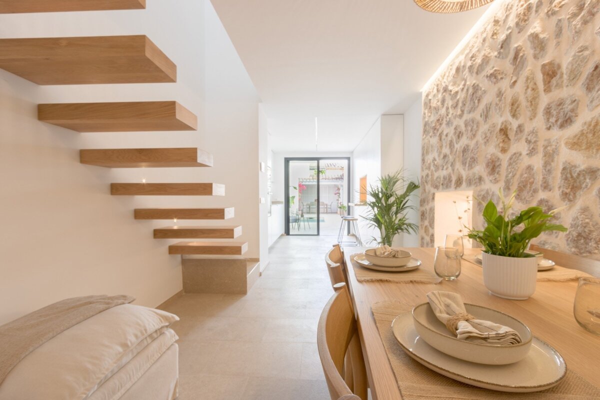 A modern dining room features a long wooden table set for guests, floating wooden stairs, and a rustic stone accent wall.