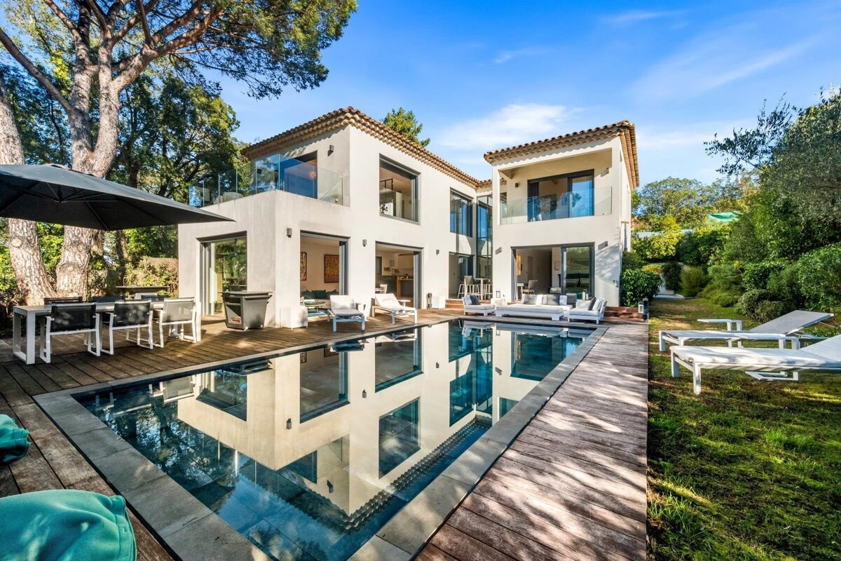 A luxurious modern white villa with a sparkling swimming pool, wooden deck, and outdoor lounge areas, surrounded by green trees.