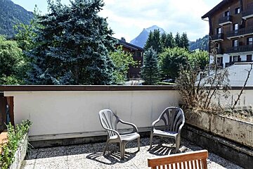 A balcony with two grey chairs offers a mountain view, dense pine trees, and distant buildings under a clear, sunny sky.