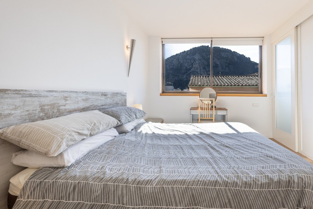 Sunlit bedroom with a gray patterned bed, rustic headboard, and a large window showcasing a dramatic mountain and tiled rooftop landscape.