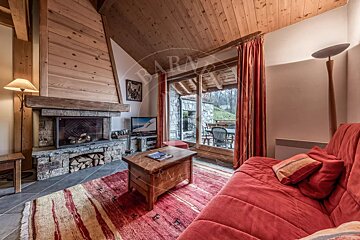Cozy, rustic living room with a large stone fireplace, red sofa, wooden ceiling, and glass doors overlooking an outdoor area.