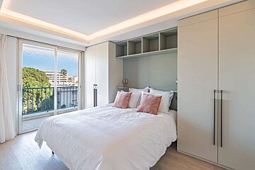 Modern bedroom with white bed, pink pillows, built-in storage, and a balcony overlooking lush trees and distant buildings.