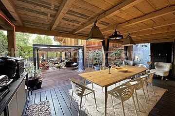 An inviting outdoor patio with a long wooden dining table, white chairs, a cozy lounge area under a pergola, and an outdoor kitchen.