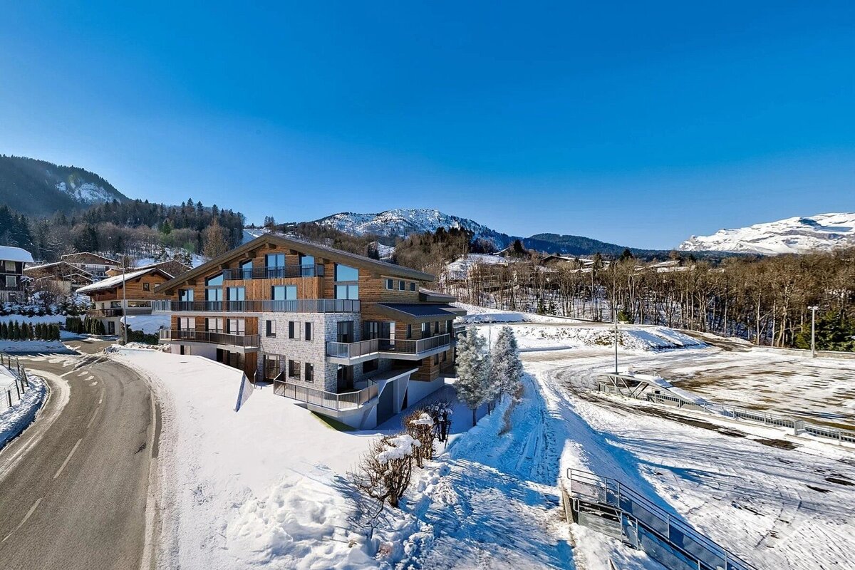 Modern wooden and stone building surrounded by snow in a sunny mountain landscape with a clear blue sky and winding road.