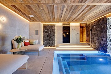Luxurious indoor pool area with a rustic wooden ceiling, stone accent walls, plush loungers, and modern ambient lighting.