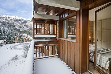A snow-covered wooden balcony in a mountain chalet, offering scenic views of a snowy village and mountains. A peek inside a cozy bedroom is visible.