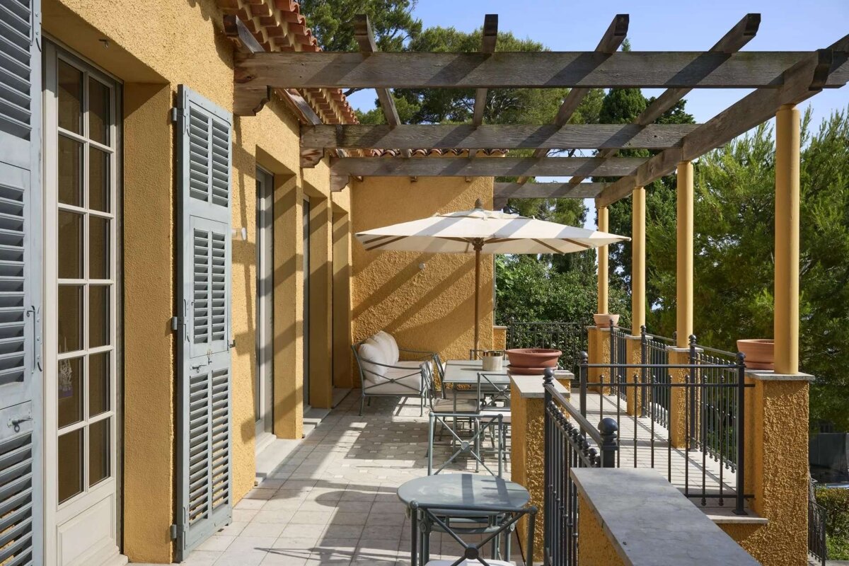 A sunlit Mediterranean terrace with a yellow stucco building, wooden pergola, patio furniture under an umbrella, and green trees beyond.