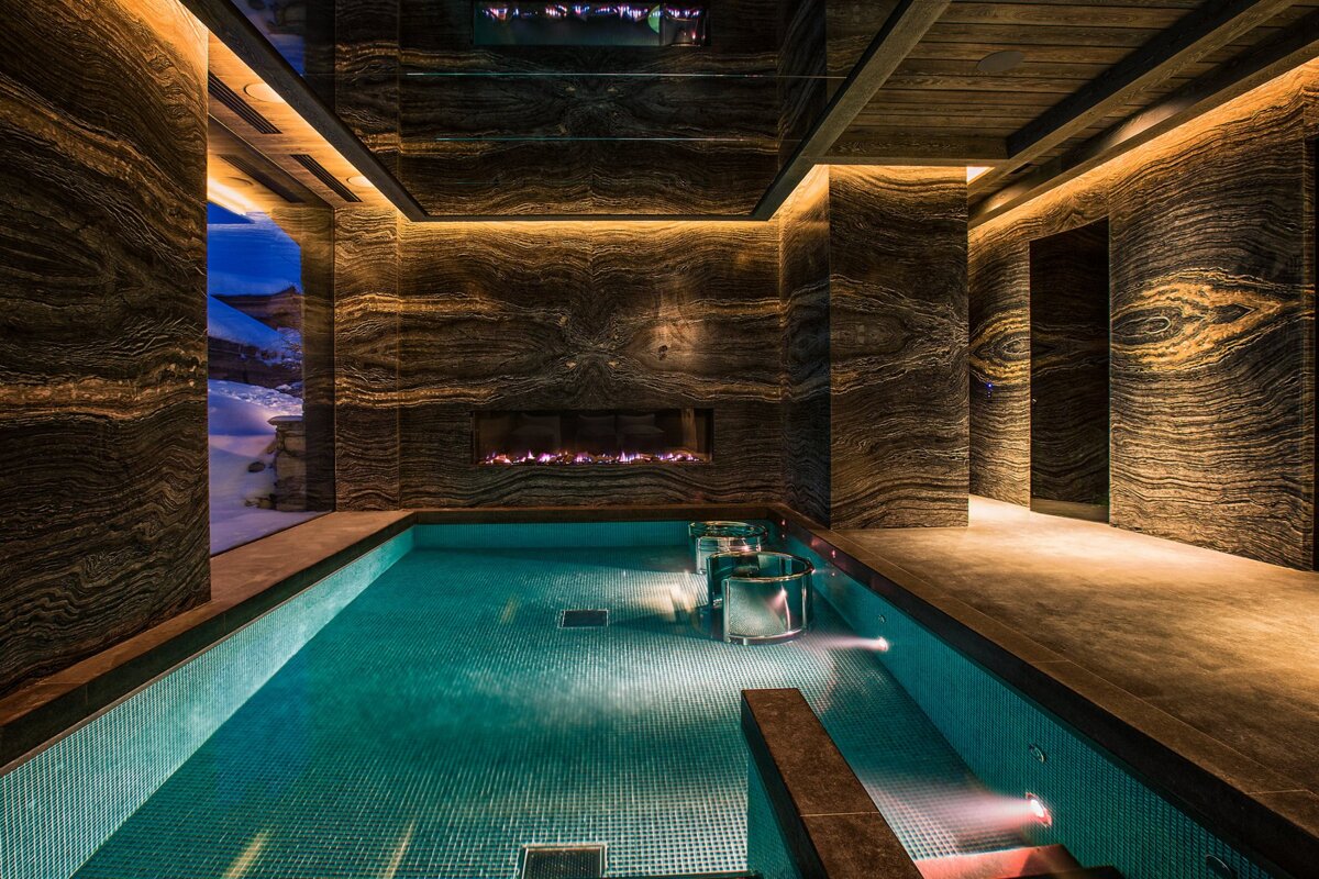 Luxurious indoor pool with patterned marble walls, a glowing fireplace, warm lighting, and a view of snowy outdoors.