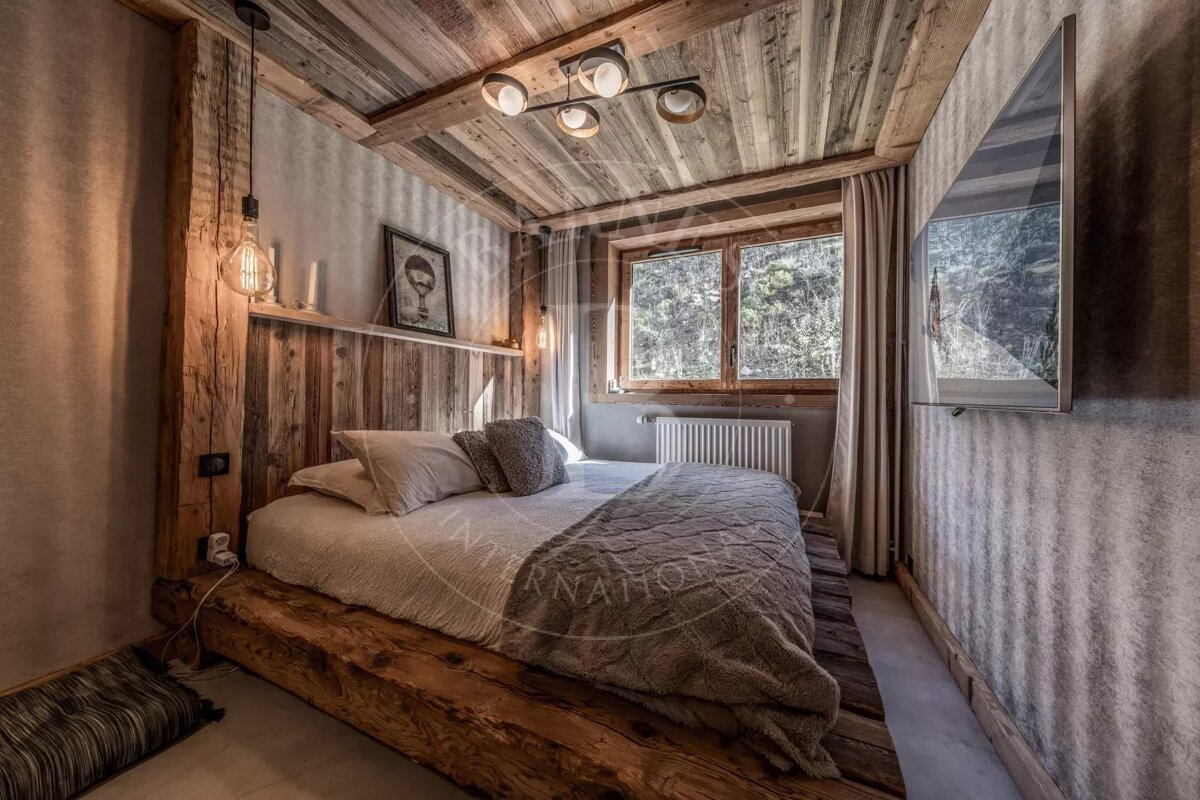 A rustic, cozy bedroom with wooden plank walls and ceiling, a large bed, warm lighting, and a window overlooking trees outside.