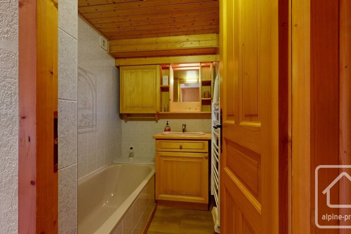 A warm, chalet-style bathroom featuring a wooden ceiling, vanity, and mirror cabinet. It includes a bathtub, sink, and white tiled walls.