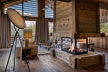 Cozy, wood-filled chalet living room with a modern glass fireplace, plush sofa, unique lamp, and large windows offering a snowy mountain vista.