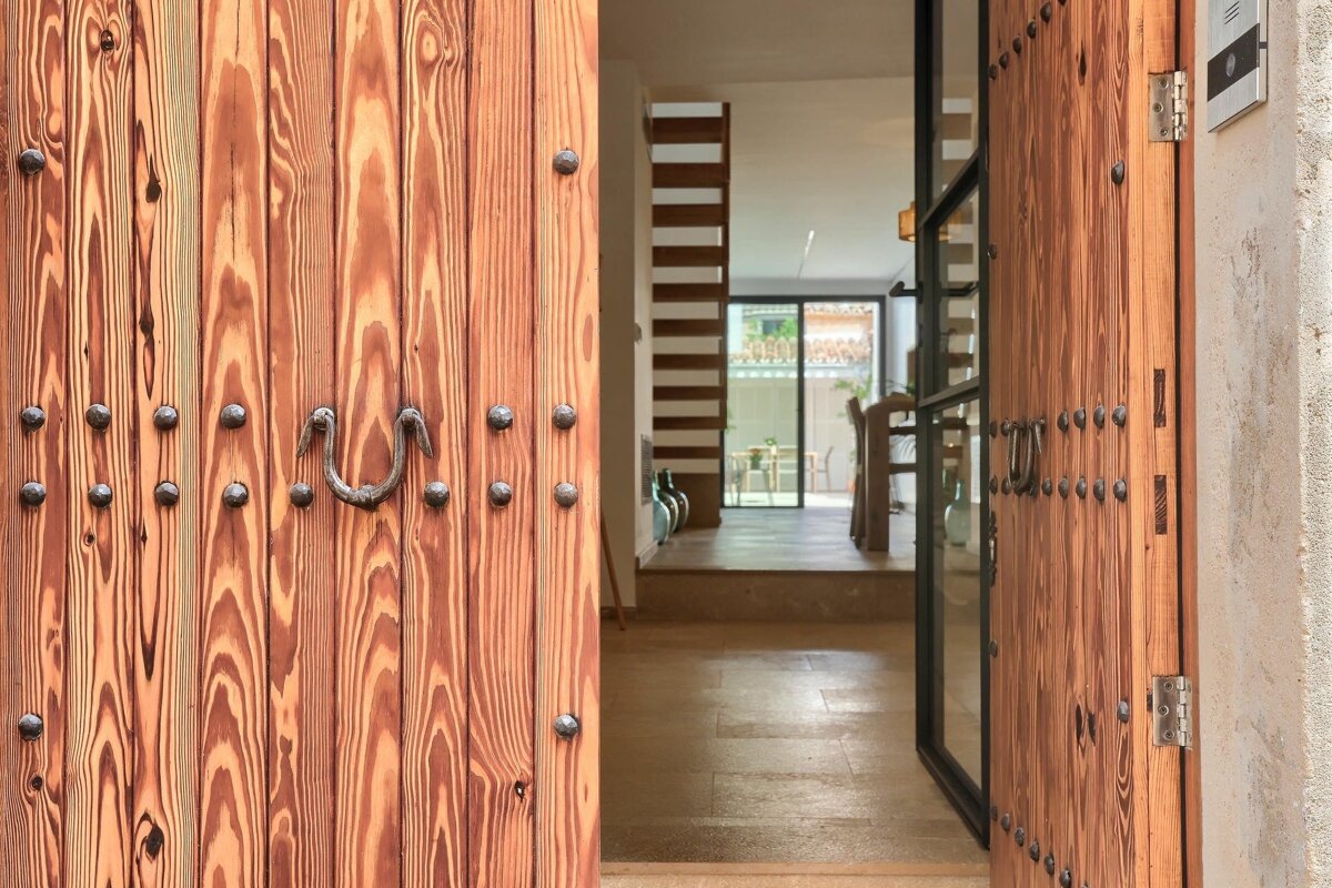 An open, rustic wooden door with metal studs and a handle reveals a bright, modern home interior with a dining area and outdoor view.