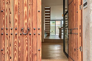 An open rustic wooden door, adorned with metal studs and a handle, reveals a bright, modern home interior with tiled floors and a dining area.