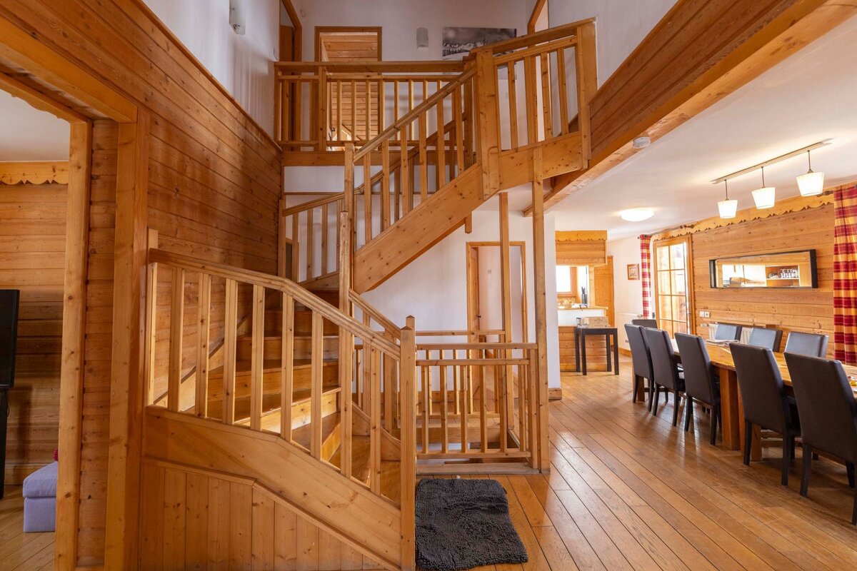 A cozy, wood-paneled lodge interior showcasing a prominent multi-level wooden staircase leading to upper floors, with a dining area on the right.
